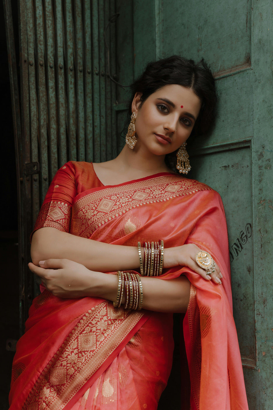 A woman in a red silk saree with a gold woven border, arms folded