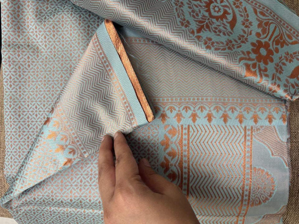 A hand turning back the woven border of a pale blue and copper silk, showing the pattern on both sides